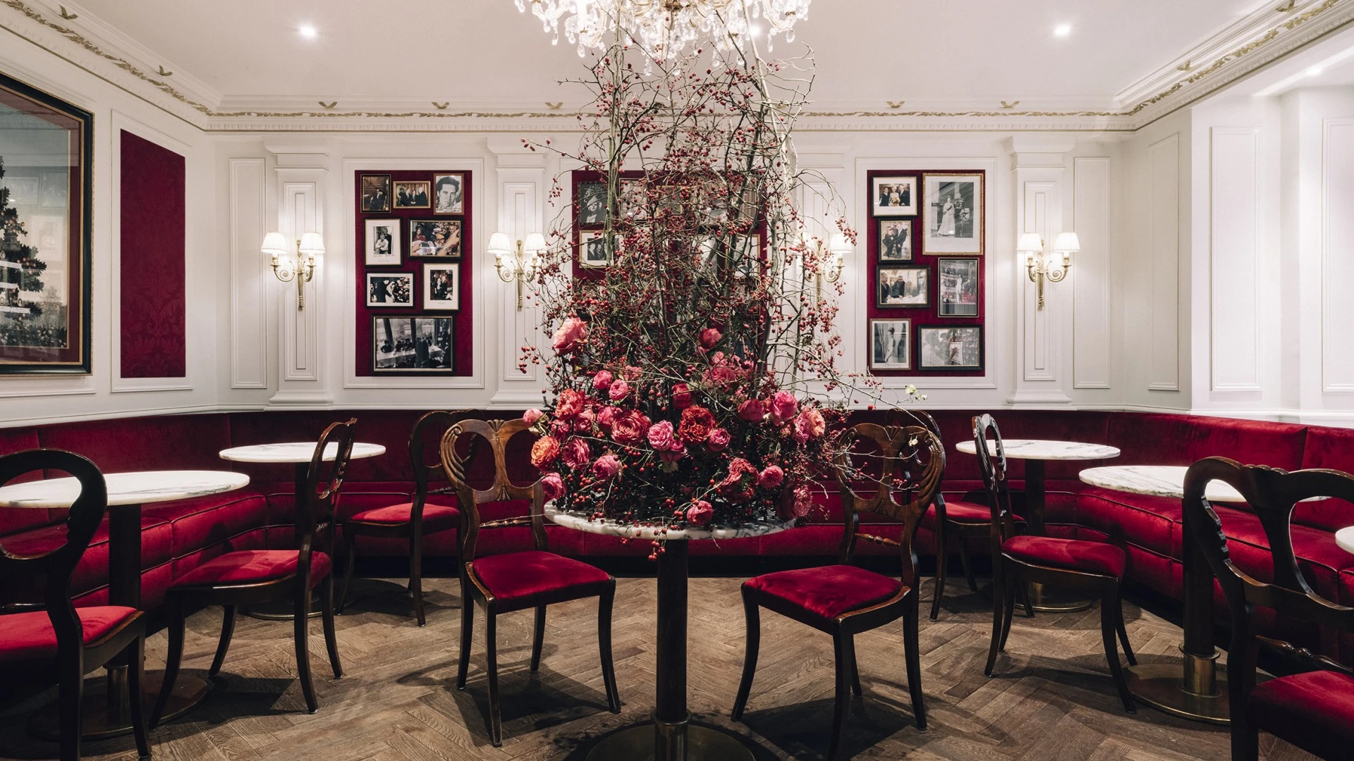 Ein stilvolles Kaffeehaus mit eleganten Marmortischen und samtroten Sitzbänken. Im Zentrum steht ein kunstvoll arrangiertes Blumenbouquet in Rosa- und Rottönen. Der Raum ist mit goldenen Wandlampen, weißen Wandvertäfelungen und gerahmten Fotografien dekoriert. Ein großer Kronleuchter rundet das edle Ambiente ab.