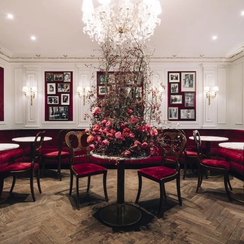 Ein stilvolles Kaffeehaus mit eleganten Marmortischen und samtroten Sitzbänken. Im Zentrum steht ein kunstvoll arrangiertes Blumenbouquet in Rosa- und Rottönen. Der Raum ist mit goldenen Wandlampen, weißen Wandvertäfelungen und gerahmten Fotografien dekoriert. Ein großer Kronleuchter rundet das edle Ambiente ab.