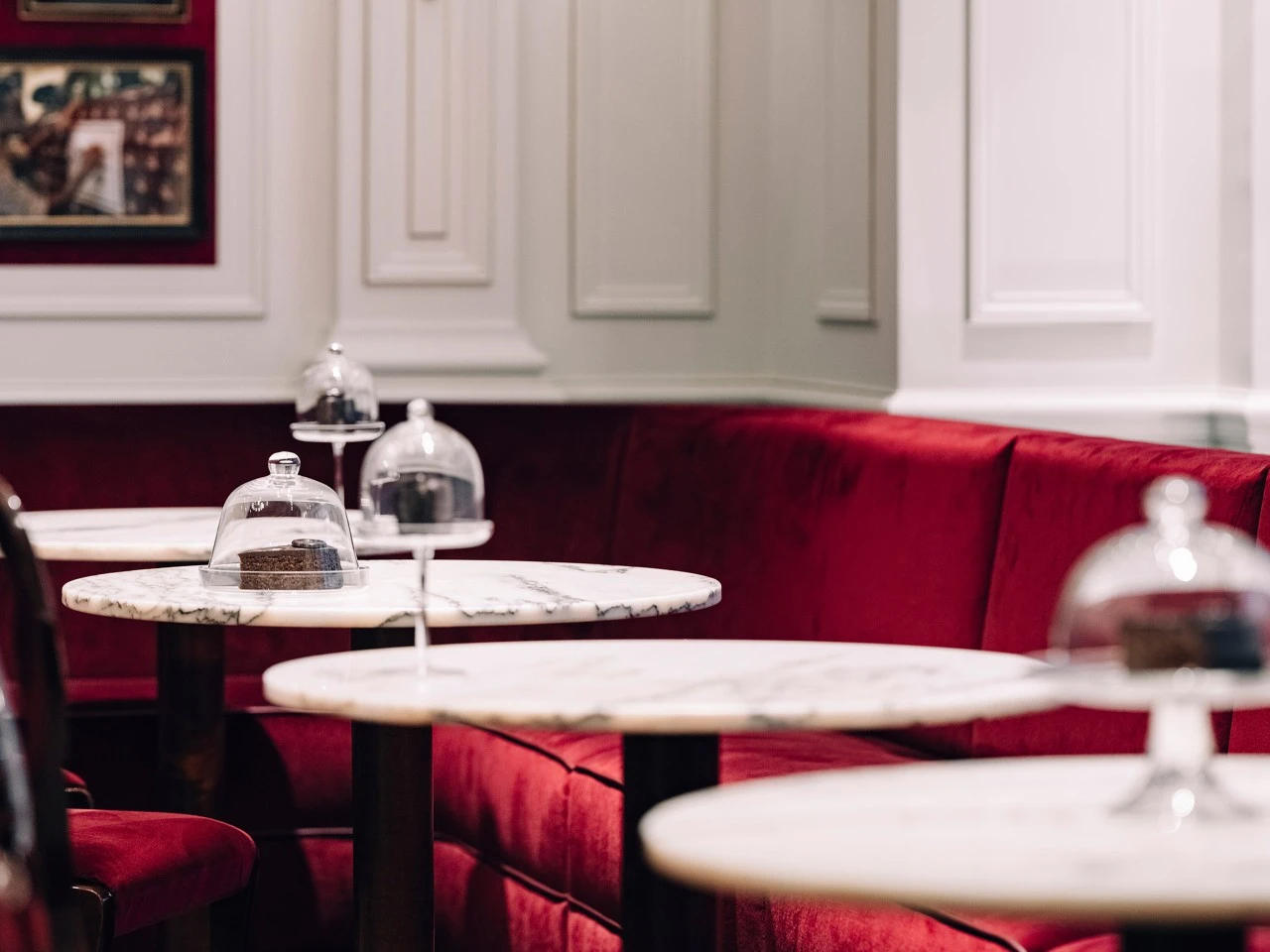 Mehrere runde Marmortische in Café Sacher Salzburg mit roten Samtbänken, darauf Original Sacher-Torten unter Glasglocken