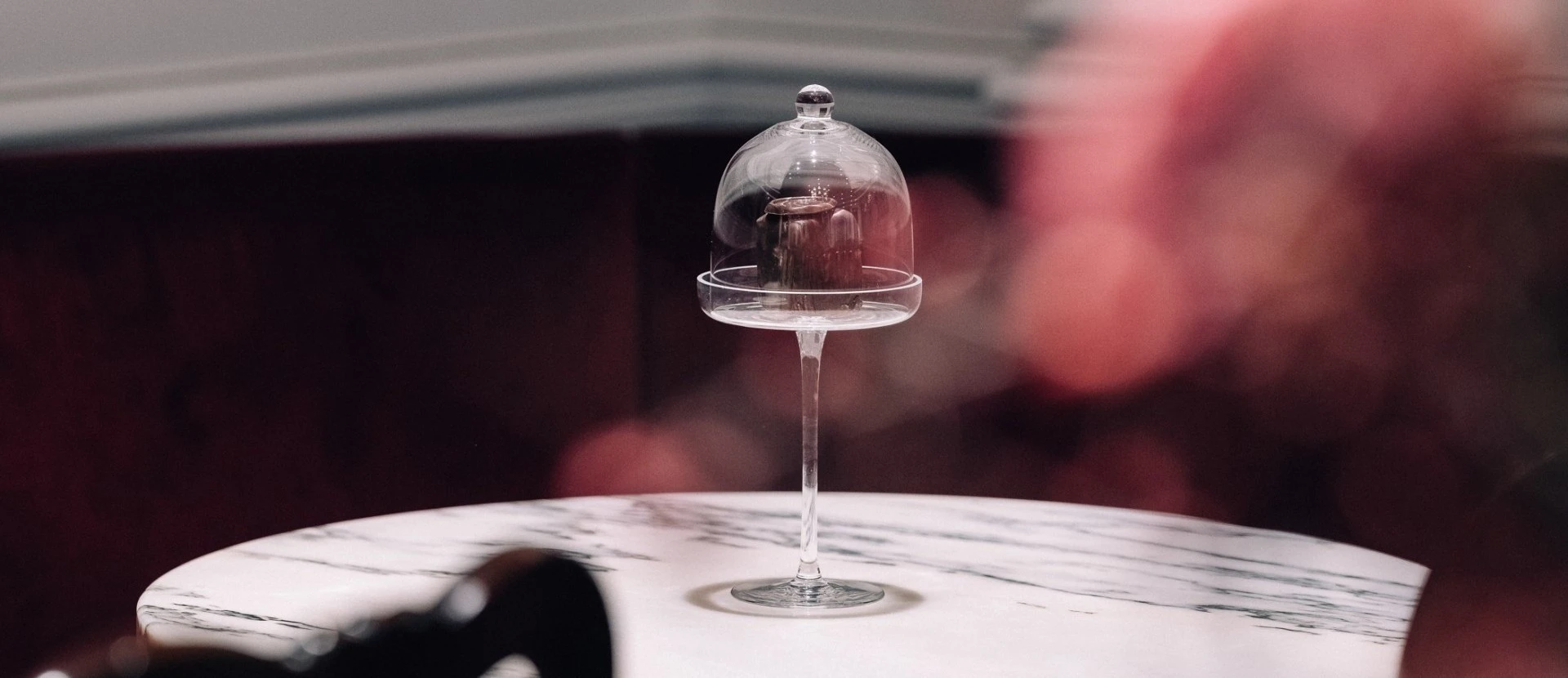 Ein einzelner Original Sacher-Würfel steht elegant auf einem hohen Glasfuß unter einer gläsernen Glocke. Der Tisch aus weißem Marmor verleiht dem Arrangement zusätzliche Raffinesse, im Hintergrund sorgt ein roter Samtton für eine warme, luxuriöse Atmosphäre.