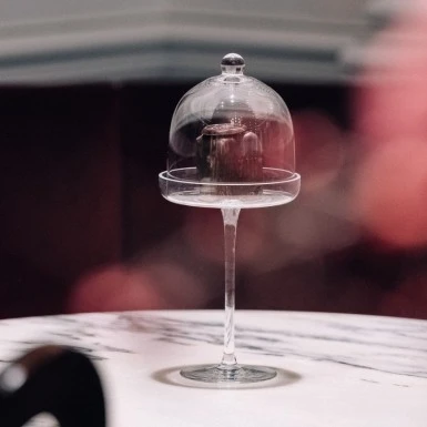 Ein einzelner Original Sacher-Würfel steht elegant auf einem hohen Glasfuß unter einer gläsernen Glocke. Der Tisch aus weißem Marmor verleiht dem Arrangement zusätzliche Raffinesse, im Hintergrund sorgt ein roter Samtton für eine warme, luxuriöse Atmosphäre.