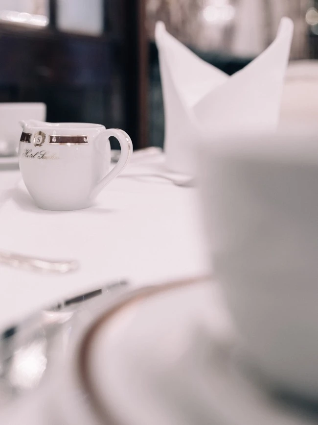 Elegant gedeckter Tisch mit einer kleinen weißen Porzellankanne des Hotel Sacher. Im Hintergrund eine kunstvoll gefaltete Stoffserviette und im Vordergrund unscharf ein Teller mit silbernem Rand.