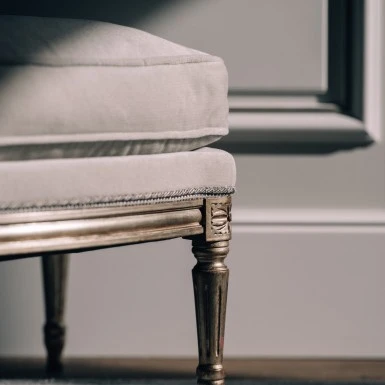 Detailaufnahme eines eleganten Möbelstücks mit vergoldetem Holzbein und heller Polsterung auf einem Teppichboden. Im Hintergrund sind weiße, klassisch verzierte Wandpaneele zu erkennen.