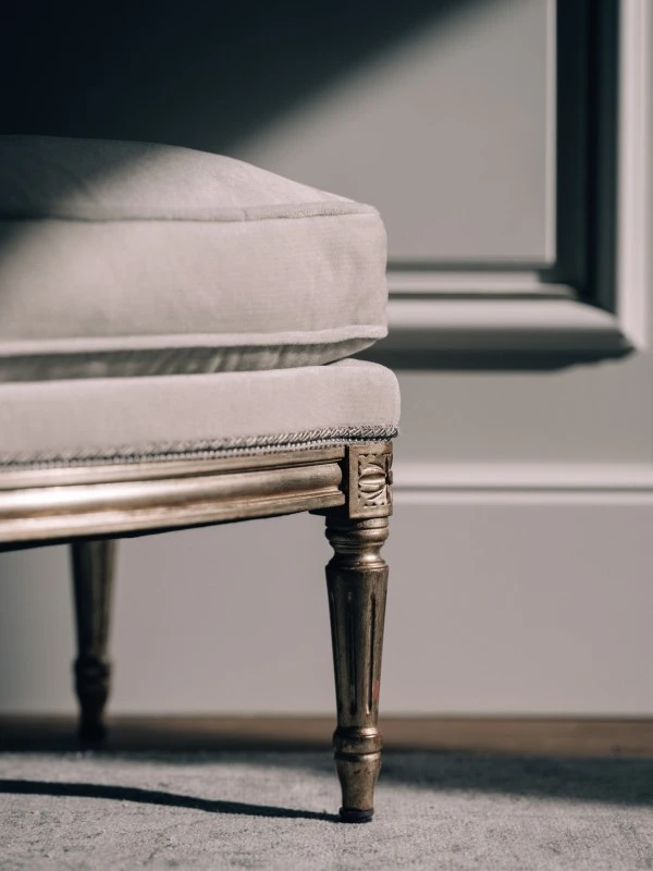 Detailaufnahme eines eleganten Möbelstücks mit vergoldetem Holzbein und heller Polsterung auf einem Teppichboden. Im Hintergrund sind weiße, klassisch verzierte Wandpaneele zu erkennen.