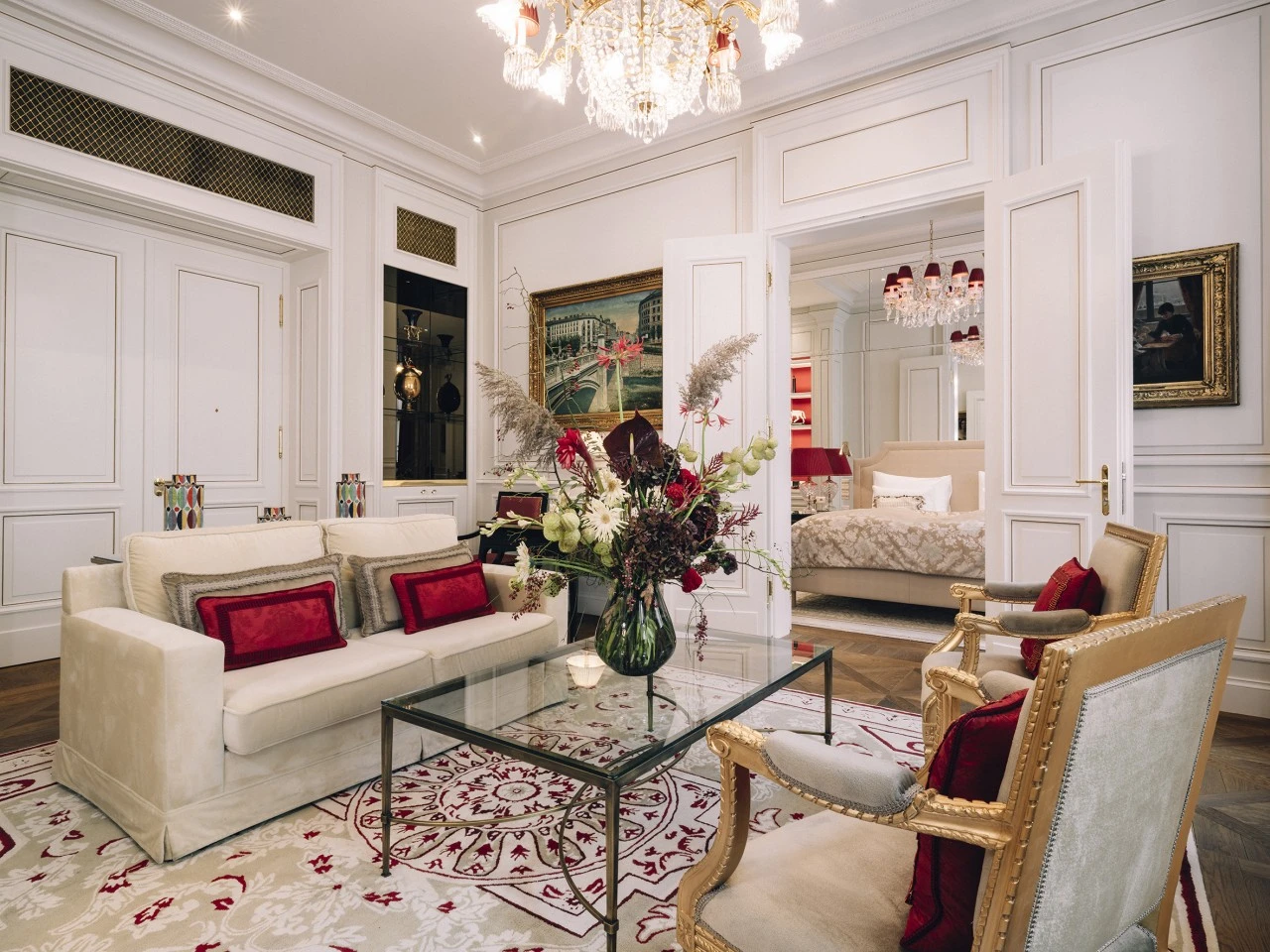 Eleganter Wohnbereich mit cremefarbenem Sofa und goldenen Sesseln mit roten Akzenten. In der Mitte steht ein Glastisch mit einem großen Blumenarrangement. Der Raum ist hell gestaltet mit weißen Wänden, Stuckverzierungen und einem Kronleuchter. Im Hintergrund ist ein Schlafzimmer sichtbar.