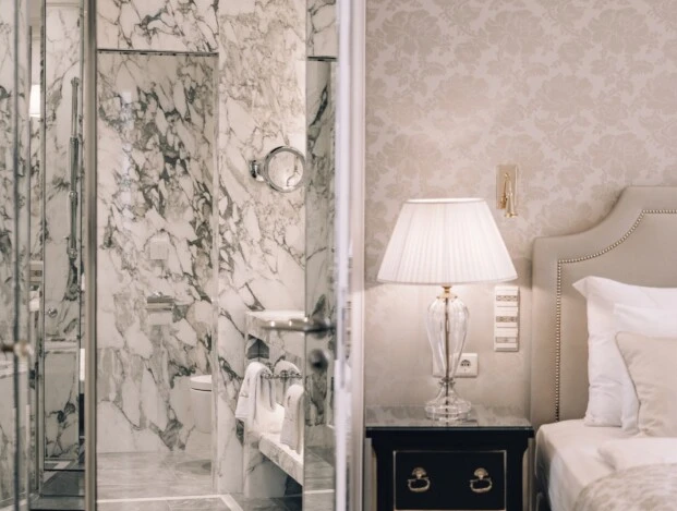 Ein elegantes Schlafzimmer mit Blick auf ein luxuriöses Badezimmer. Neben dem Bett steht ein schwarzer Nachttisch mit einer edlen Glaslampe. Das Badezimmer beeindruckt mit hellem Marmor und glänzenden Armaturen.