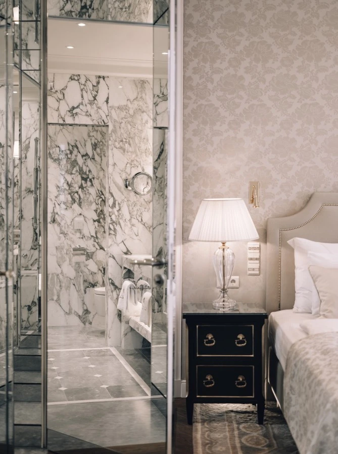 Ein elegantes Schlafzimmer mit Blick auf ein luxuriöses Badezimmer. Neben dem Bett steht ein schwarzer Nachttisch mit einer edlen Glaslampe. Das Badezimmer beeindruckt mit hellem Marmor und glänzenden Armaturen.