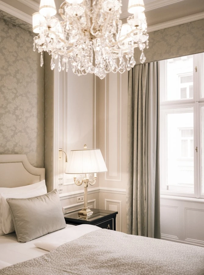 Hotel Sacher Wien, Suite Ein stilvoll eingerichtetes Schlafzimmer mit einem gepolsterten Bett, einer eleganten Nachttischlampe und einem funkelnden Kronleuchter. Durch das Fenster fällt weiches Tageslicht in den Raum mit klassischer Wandvertäfelung und floralen Tapeten.