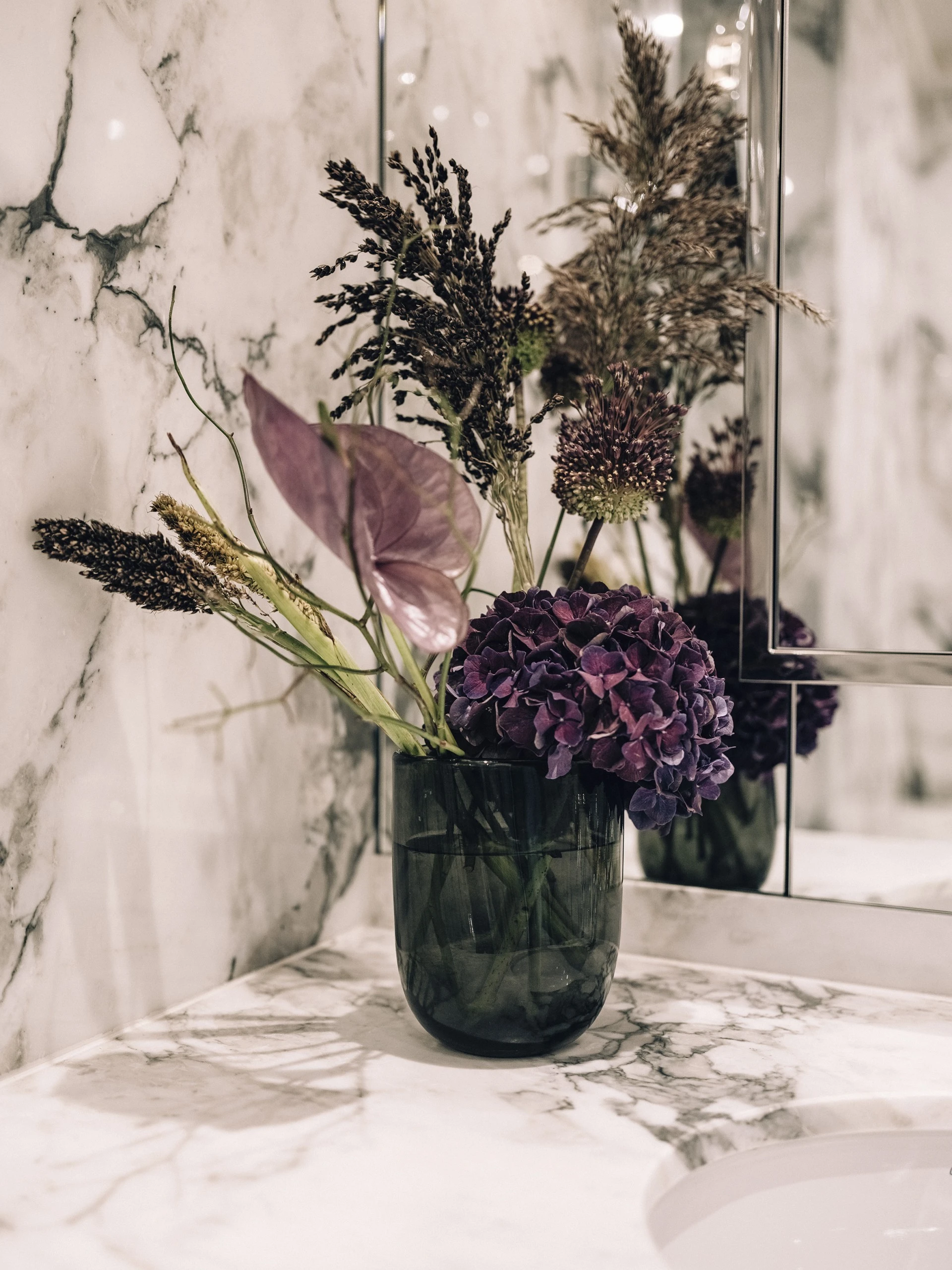 Ein Blumenarrangement in einem dunklen Glasgefäß auf einer Marmorablage. Die Blumen bestehen aus verschiedenen getrockneten und frischen Elementen in Violett- und Naturtönen.