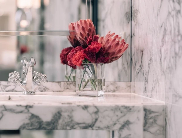Ein elegantes Waschbecken aus weißem Marmor mit verchromtem Wasserhahn, davor ein Glas mit roten Blumen, darunter ein flauschiges Handtuch mit gesticktem Emblem auf einer verchromten Halterung. Im Hintergrund Spiegel und ein runder Kosmetikspiegel.