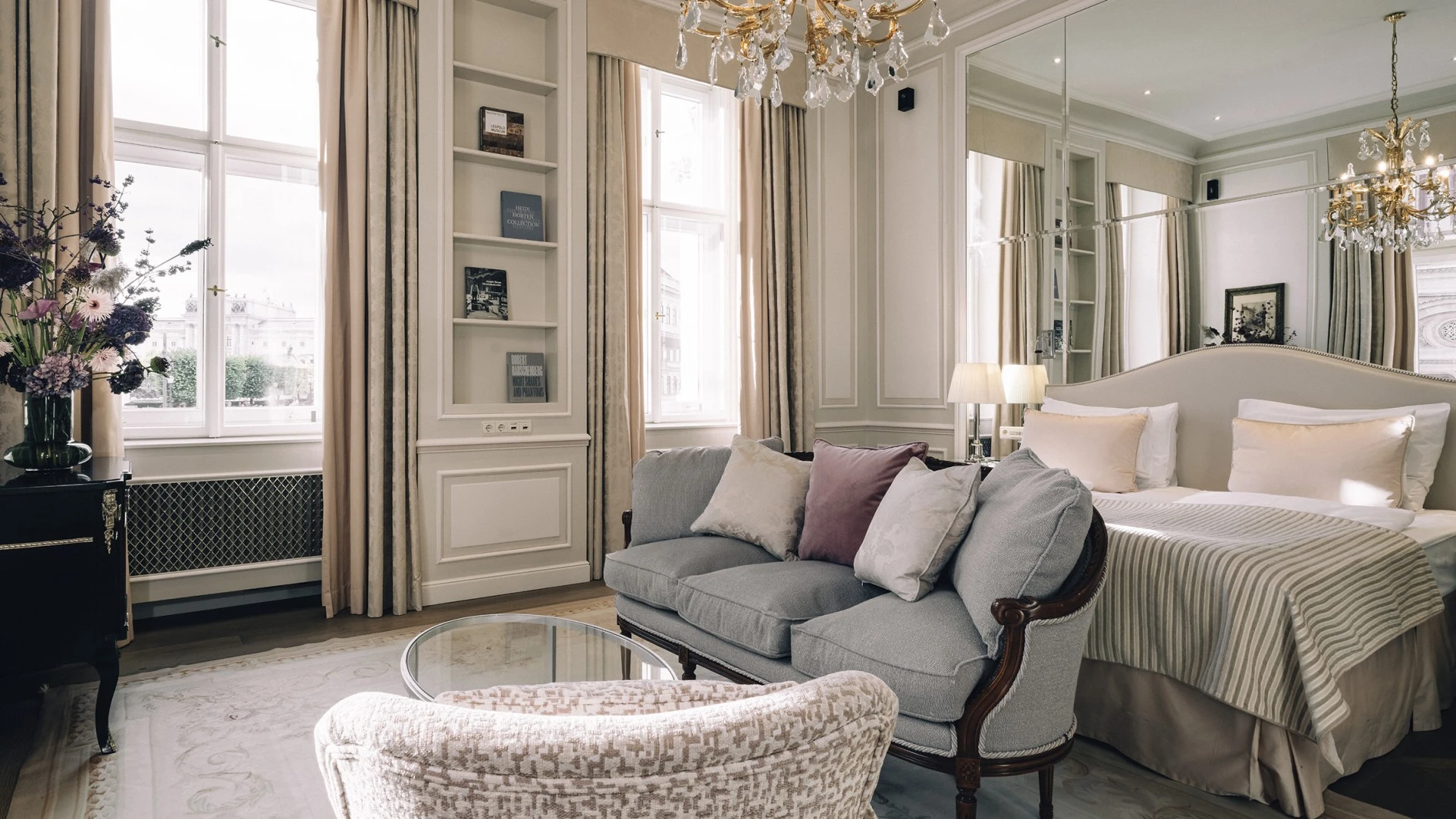 Ein elegantes Schlafzimmer mit großem Spiegel hinter dem Bett, zwei Kronleuchtern, einem Himmelbett mit gestreifter Tagesdecke sowie einer kleinen Sitzecke mit Sofa, Sessel und Glastisch. Links ein Fenster mit Blick auf ein klassizistisches Gebäude und ein Blumenarrangement auf einer Kommode.