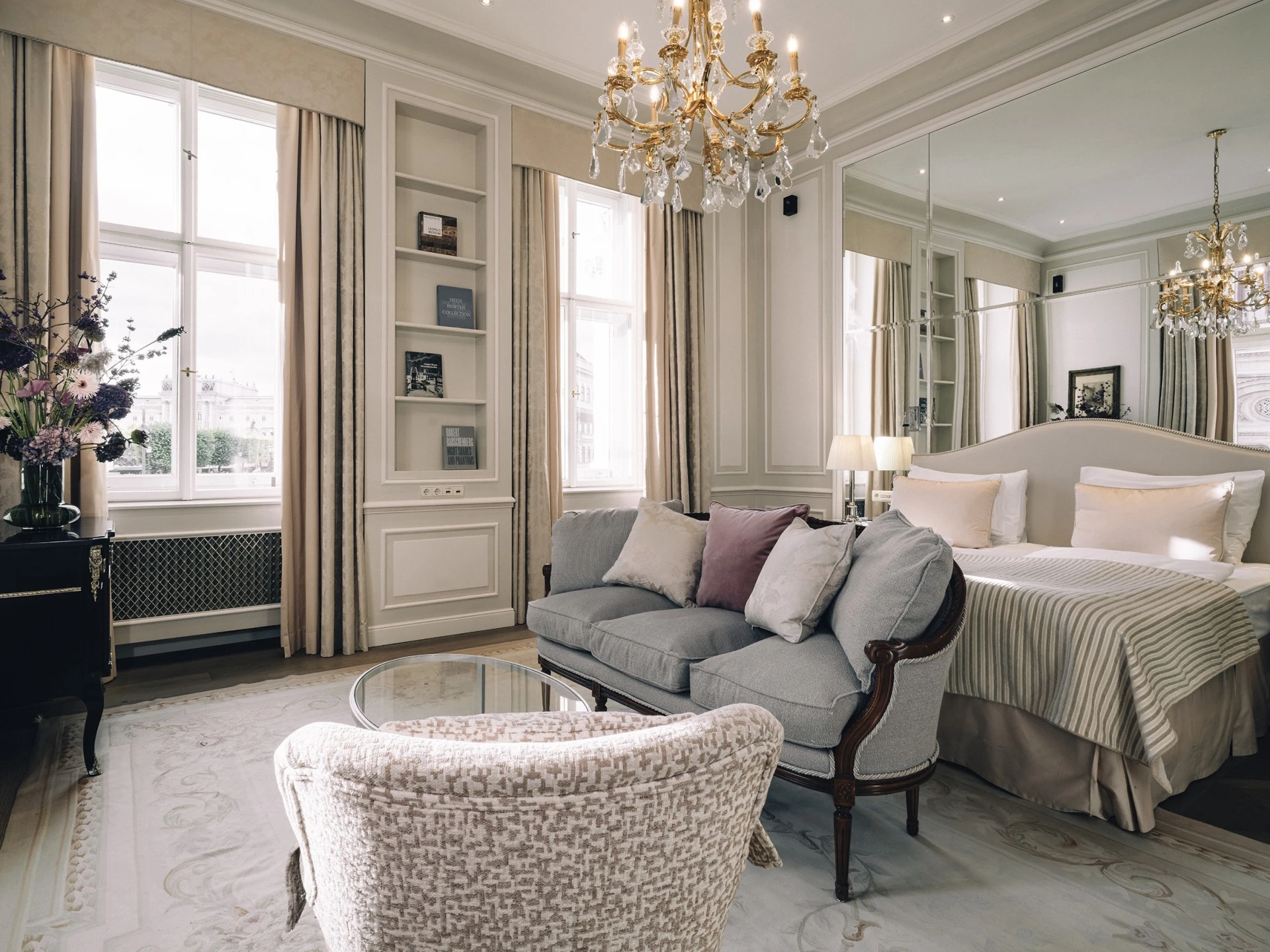 Ein elegantes Schlafzimmer mit großem Spiegel hinter dem Bett, zwei Kronleuchtern, einem Himmelbett mit gestreifter Tagesdecke sowie einer kleinen Sitzecke mit Sofa, Sessel und Glastisch. Links ein Fenster mit Blick auf ein klassizistisches Gebäude und ein Blumenarrangement auf einer Kommode.