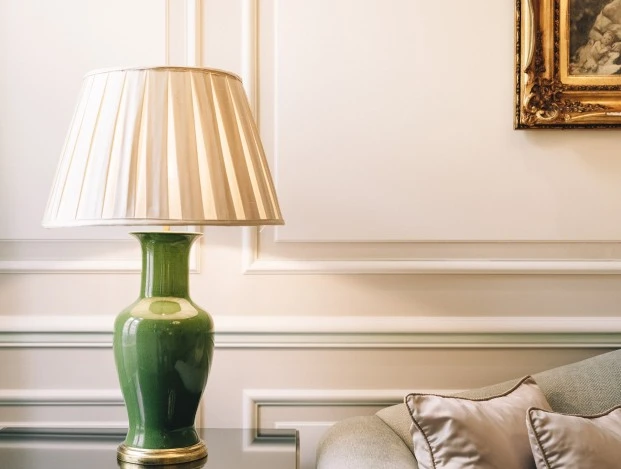 Hotel Sacher Wien, Suite Ein eleganter Ausschnitt eines Wohnbereichs mit einer auffälligen grünen Tischlampe auf einem schwarzen Beistelltisch mit Glasplatte. Daneben eine hellgraue Couch mit glänzenden, champagnerfarbenen Kissen. Die Wand ist klassisch mit Stuckleisten verziert, und ein goldgerahmtes Gemälde sorgt für einen kunstvollen Akzent im Hintergrund.