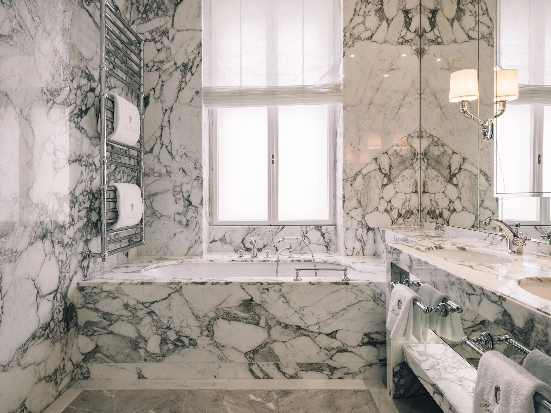 Luxuriöses Badezimmer mit edlem weißen Marmor und grauer Maserung. Badewanne und Waschtisch sind in Marmor eingefasst, dazu verchromte Armaturen, Handtuchhalter und warme Lichtakzente.