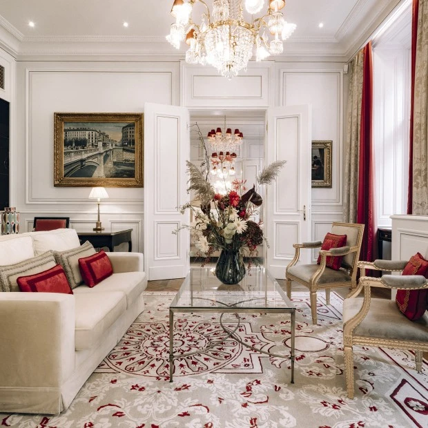 Elegantes Hotelzimmer mit Kingsize-Bett, luxuriösem Teppich und gemütlicher Sitzecke. Große Fenster mit schweren Vorhängen lassen viel Tageslicht hinein. Kristalllüster und klassische Gemälde runden das edle Ambiente ab.