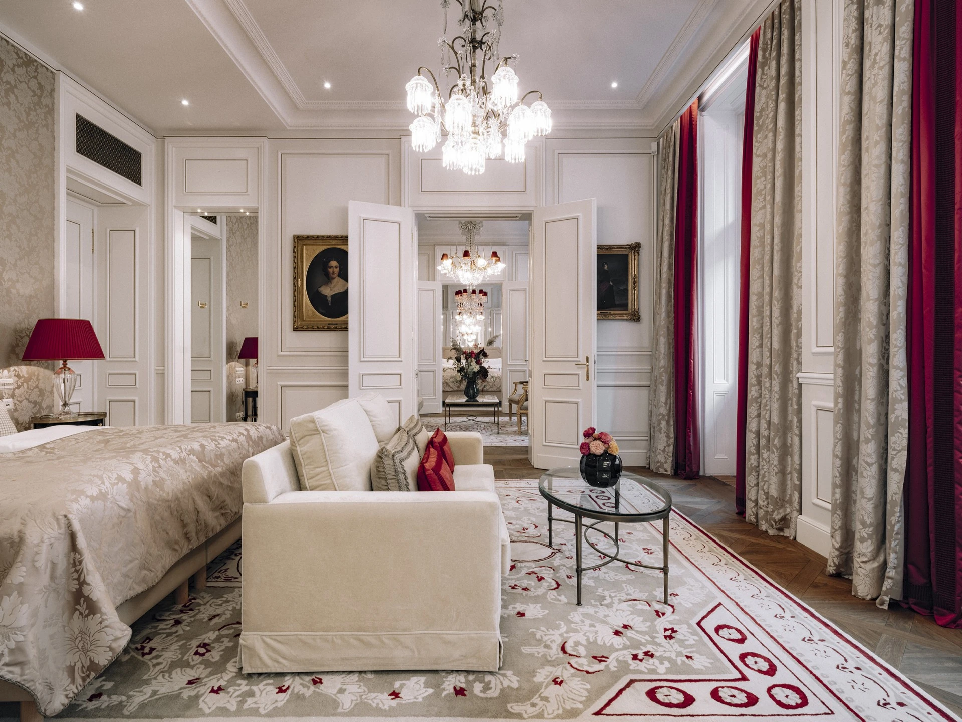 Elegantes Hotelzimmer mit Kingsize-Bett, luxuriösem Teppich und gemütlicher Sitzecke. Große Fenster mit schweren Vorhängen lassen viel Tageslicht hinein. Kristalllüster und klassische Gemälde runden das edle Ambiente ab.