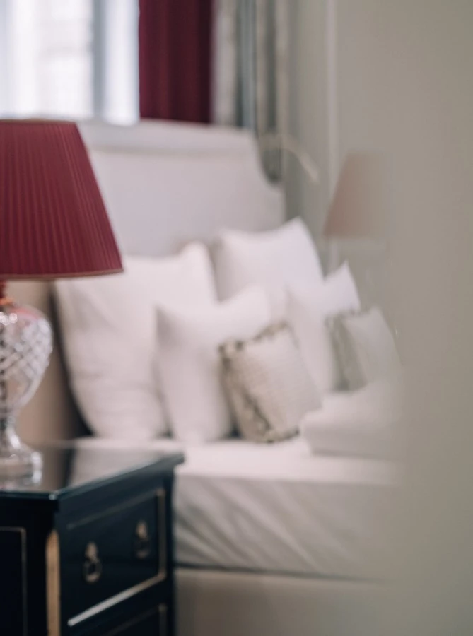 Ein Nachttisch mit dunkler Hochglanzoberfläche und goldenen Details steht neben einem Bett mit hellem Polsterkopfteil. Darauf steht eine Tischlampe mit klarem Kristallfuß und einem rot gefalteten Lampenschirm. Das Bett ist mit mehreren weißen Kissen und einem kleinen Zierkissen in Rautenstruktur arrangiert.
