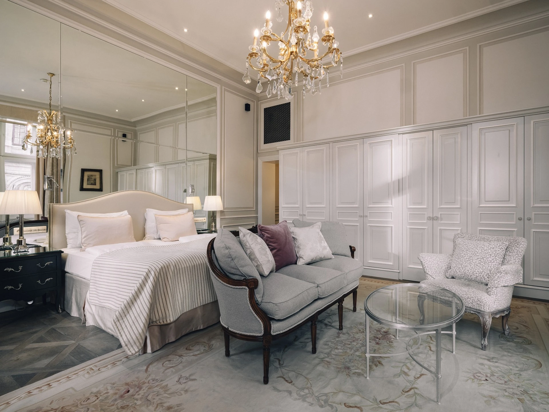 Das Zimmer ist mit einem großen, gepolsterten Bett vor einer wandfüllenden Spiegelfläche ausgestattet. Rechts davon stehen eine gepolsterte Sitzbank mit Zierkissen und ein Sessel mit floralem Muster. Davor befindet sich ein runder Glastisch.