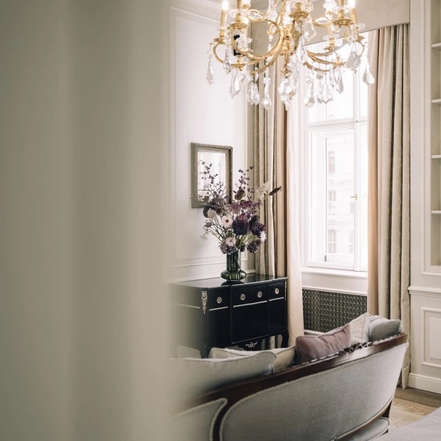Ein Kristallluster hängt von der Decke und reflektiert das Licht. Darunter steht ein schwarzes Sideboard mit silbernen Knäufen. Darauf steht eine Glasvase mit violetten, rosa und weißen Blumen. Im Vordergrund ein helles Sofa mit runden Polstern und Holzrahmen.
