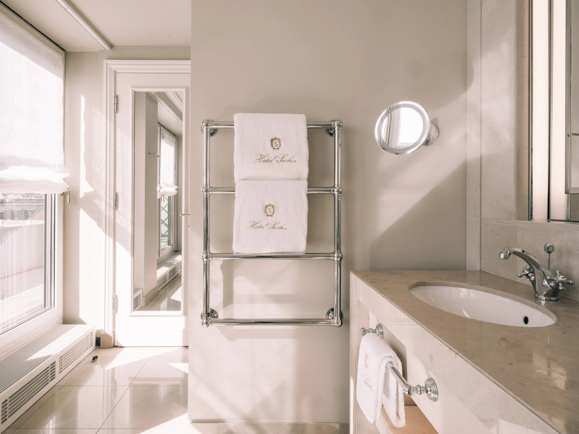 Ein helles Badezimmer mit cremefarbenen Wänden, einem Waschtisch mit beigem Marmor und eleganten Armaturen. Auf einem verchromten Handtuchhalter hängen zwei weiße Handtücher mit dem Logo des Hotel Sacher. Sonnenlicht fällt durch ein Fenster und taucht den Raum in warmes Licht.