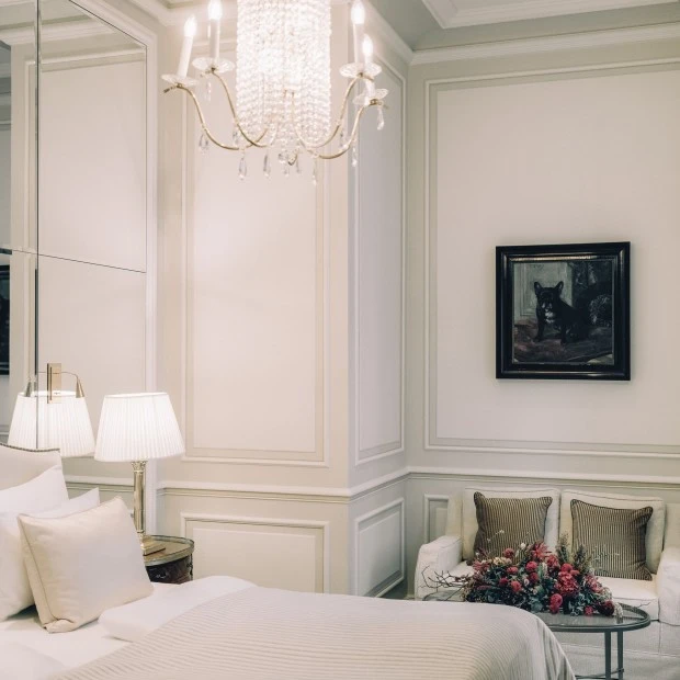 Ein Teil eines elegant eingerichteten Schlafzimmers mit einem großen Bett in heller Bettwäsche. Links steht ein Nachttisch mit einer klassischen Lampe. An der Decke hängt ein Kronleuchter mit Glasperlen und Kerzenlampen.