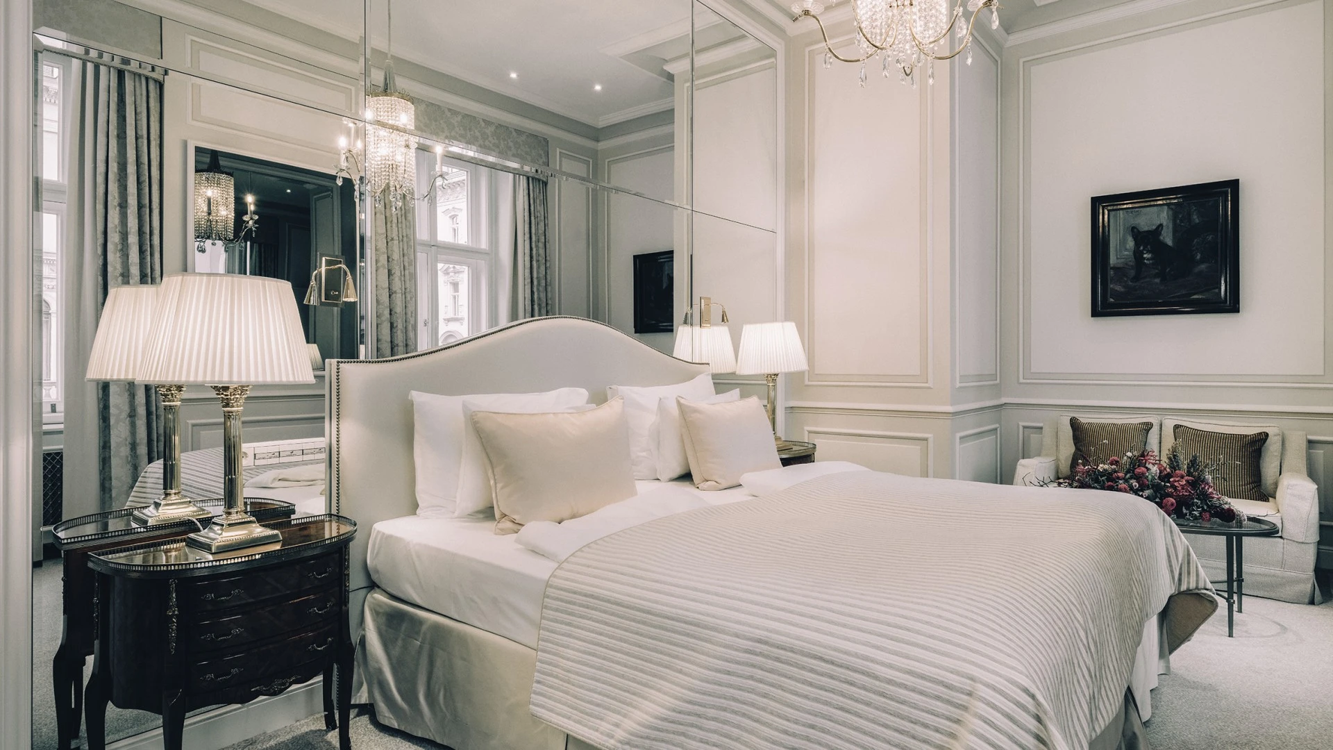 Ein großzügiges Schlafzimmer mit einem großen Bett, das mit weißer Bettwäsche und mehreren Kissen ausgestattet ist. An beiden Seiten des Betts stehen dunkle Nachttische mit vergoldeten Lampen. Hinter dem Bett erstreckt sich eine große Spiegelwand.