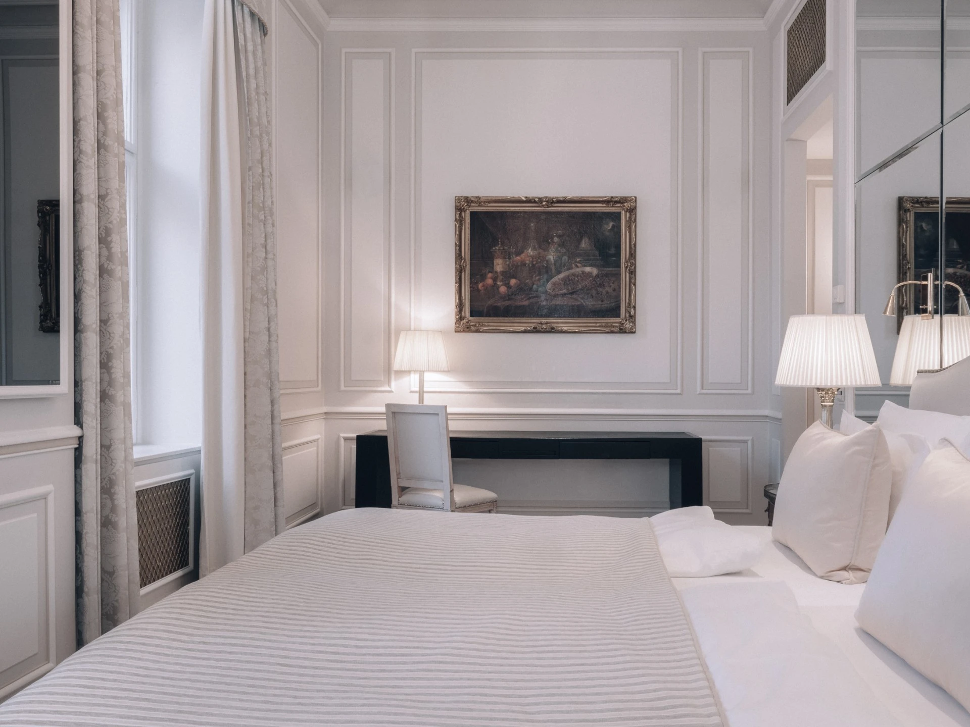 Ein elegantes Schlafzimmer mit einem großen Bett, das mit weißen Kissen und einer hell gestreiften Tagesdecke ausgestattet ist. Rechts und links neben dem Bett stehen Nachttische mit klassischen Lampen. Vor dem Fenster befindet sich ein schwarzer Schreibtisch mit einem weißen Stuhl und einer Tischlampe. An der Wand darüber hängt ein gerahmtes Gemälde mit dunklen Farbtönen. Die Wände sind mit Zierleisten verziert, die Vorhänge sind hell mit dezentem Muster.