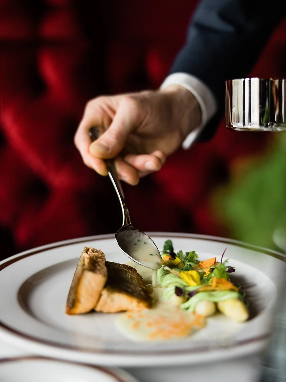 Ein Teller mit gebratenem Fischfilet, Spargel und feinen Gemüsedekorationen wird mit einer hellen Sauce verfeinert. Die Hand einer Person in dunklem Anzug reicht von oben ins Bild und träufelt die Sauce mit einem Löffel über das Gericht.