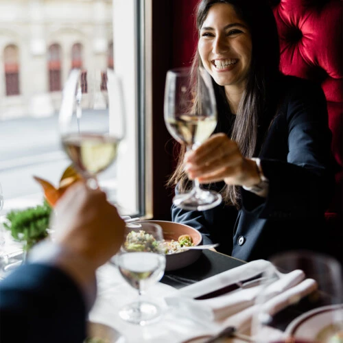 Eine Frau in dunklem Blazer sitzt im Café Bel Étage an einem gedeckten Tisch am Fenster und stößt mit einem Glas Weißwein an. Auf dem Tisch stehen mehrere Gläser und Teller mit Speisen, die rot gepolsterte Wand im Hintergrund ist mit Knopfheftung gestaltet. Durch das Fenster ist die historische Fassade eines Gebäudes zu sehen.