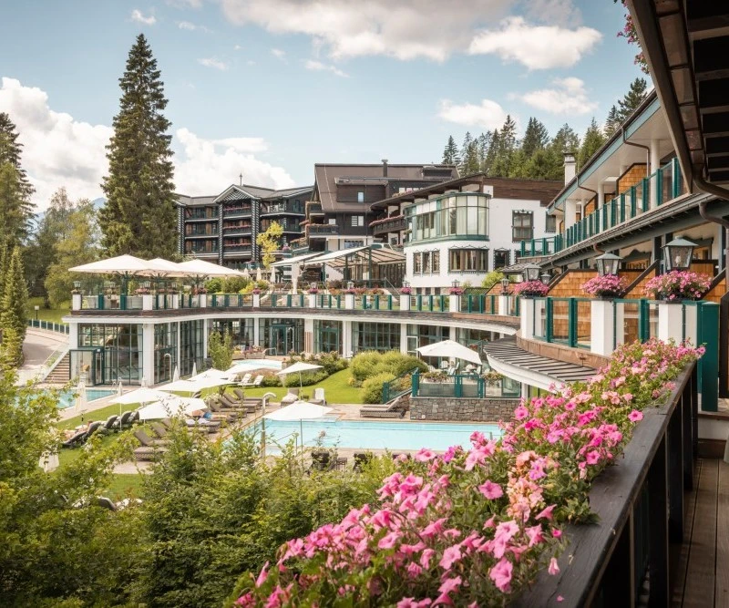Alpin Resort Sacher Außenansicht eines großen, eleganten Hotels mit mehreren Terrassen, Balkonen, bunten Blumenarrangements und einem Poolbereich, eingebettet in eine grüne Waldlandschaft.