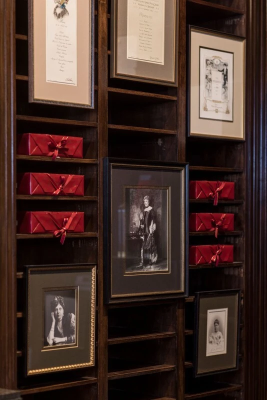 Holzwand mit integrierten Regalfächern, in denen mehrere rote Boxen mit der Original Sacher-Torte gestapelt sind. Dazwischen hängen gerahmte Schwarz-Weiß-Fotografien von Anna Sacher und anderen historischen Persönlichkeiten.