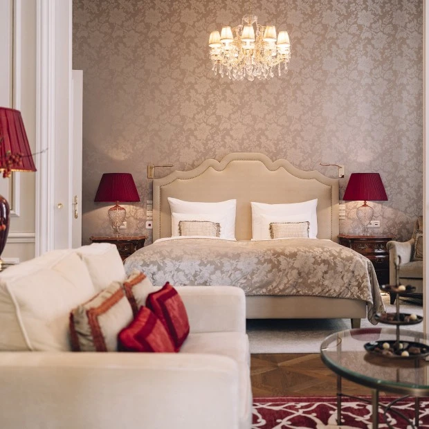Ein stilvolles Schlafzimmer mit einem eleganten, beigen Bett und edler, floraler Tapete. Warme Rottöne in Lampen und Kissen setzen Akzente. Der Kronleuchter sorgt für eine luxuriöse Atmosphäre, während klassische Möbel und Details Gemütlichkeit und Raffinesse ausstrahlen.