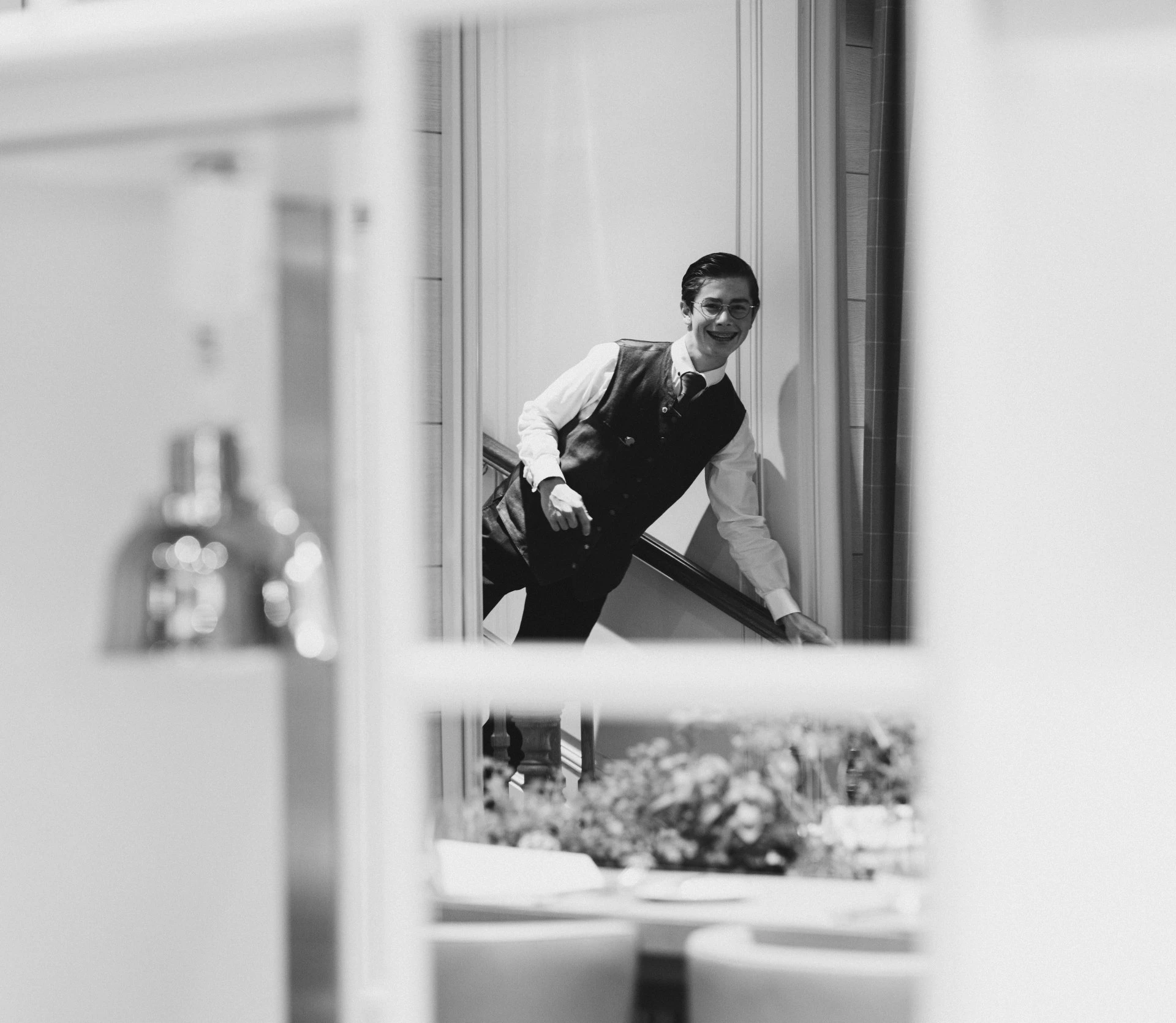 Ein Mitarbeiter in klassischer Uniform mit Weste, Hemd und Krawatte steigt lächelnd eine Treppe hinunter. Die Szene ist durch ein Fenster oder eine Öffnung fotografiert, im Vordergrund sind unscharf gedeckte Tische zu erkennen.