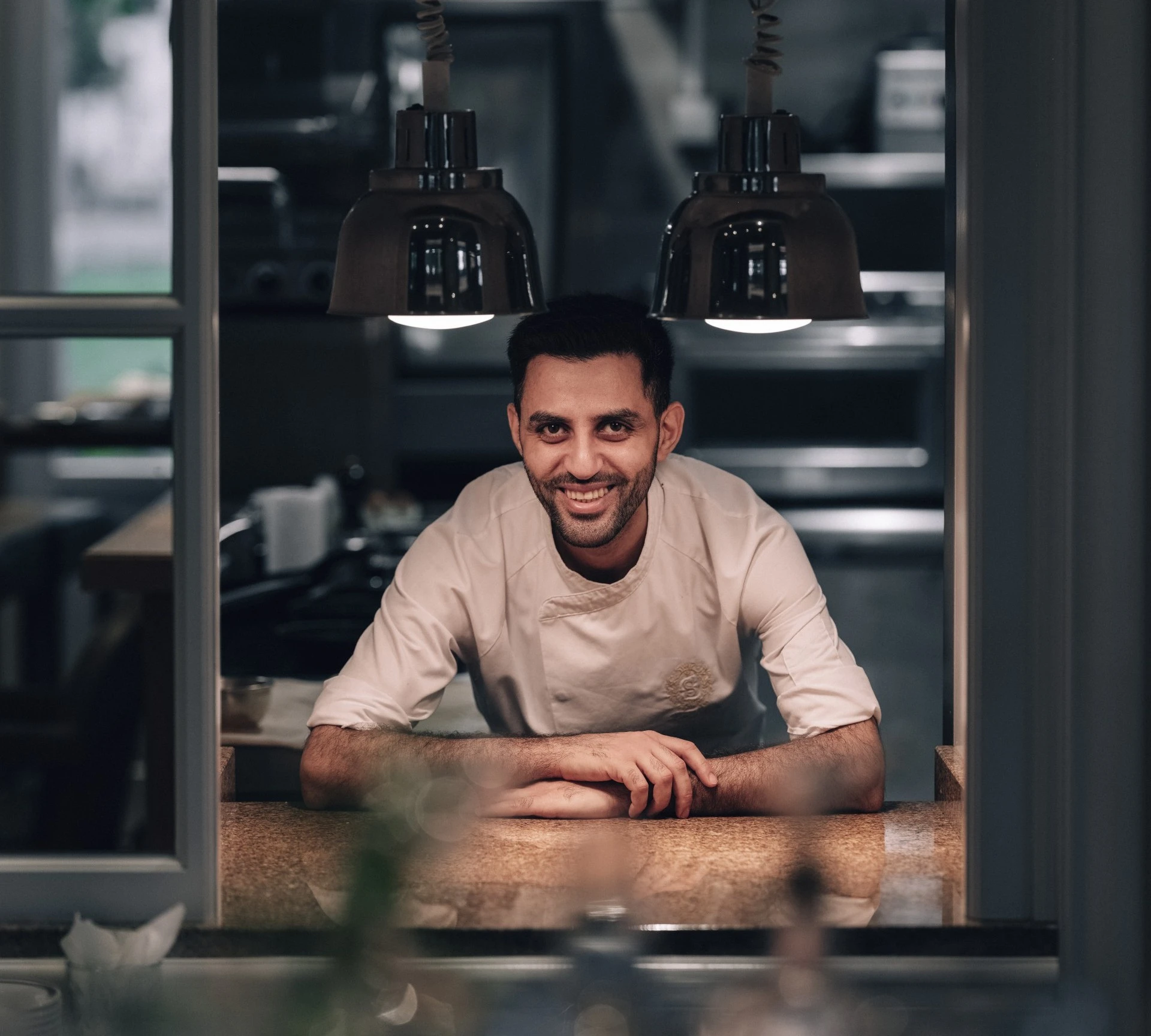 Ein Koch steht hinter der Durchreiche in einer Restaurantküche, trägt eine weiße Kochjacke mit gesticktem Sacher Emblem und lächelt freundlich in die Kamera. Die Szene ist warm beleuchtet, über ihm hängen zwei Wärmelampen.