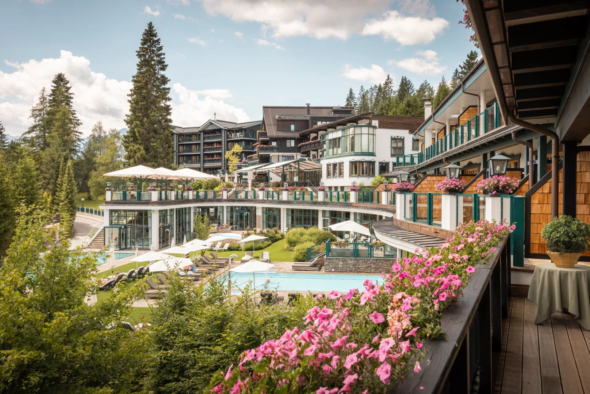 Hotelanlage in den Bergen mit mehreren Gebäudeteilen, großen Glasfronten und Balkonen mit Blumenschmuck. Im Gartenbereich befinden sich Pools, Liegen und Sonnenschirme.