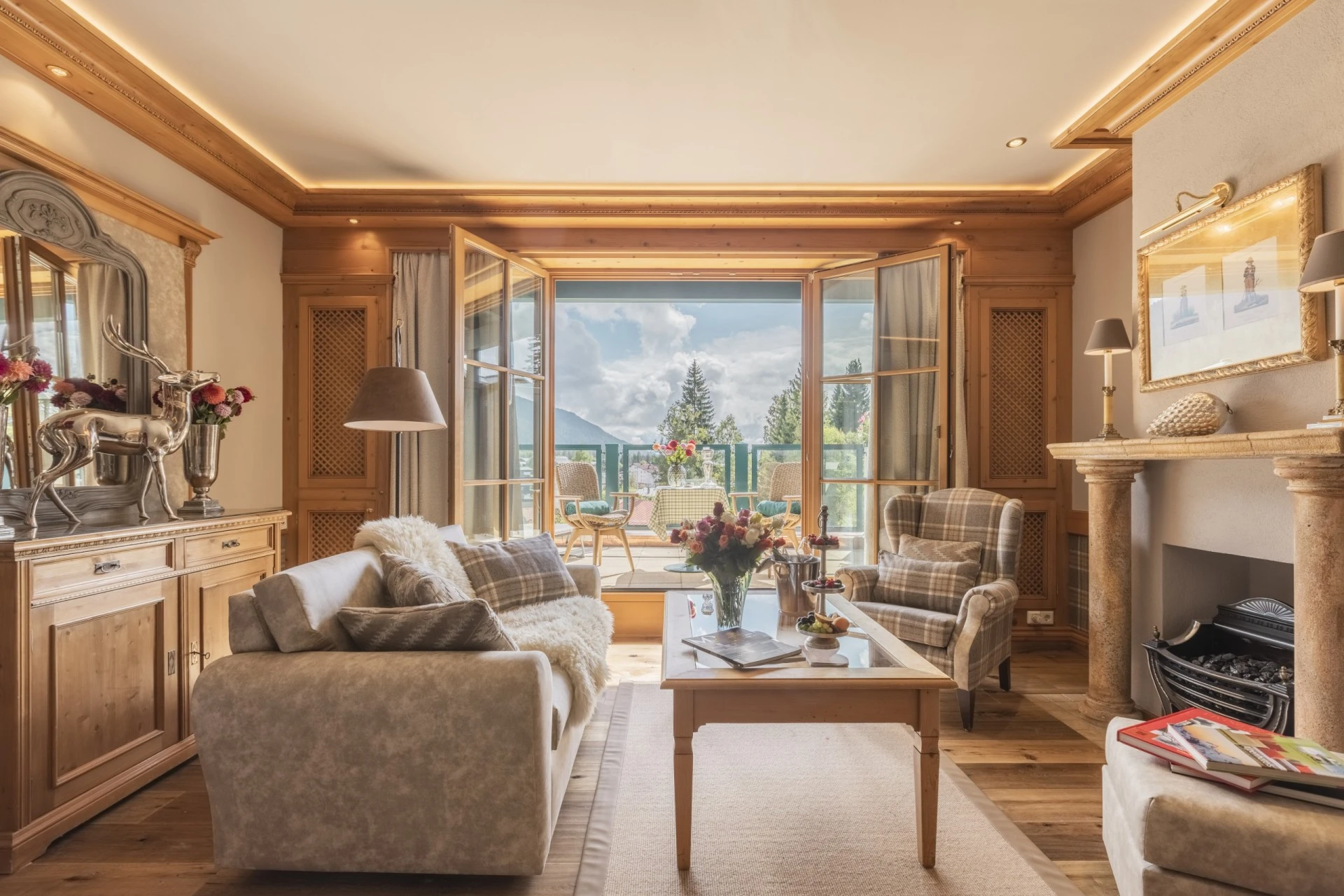 Gemütlich eingerichtetes Wohnzimmer mit Kamin, Sesseln, Sofa und offenem Zugang zur Terrasse mit Blick auf Berge und Natur. Auf dem Tisch ein Blumenstrauß, Sekt und Süßigkeiten.