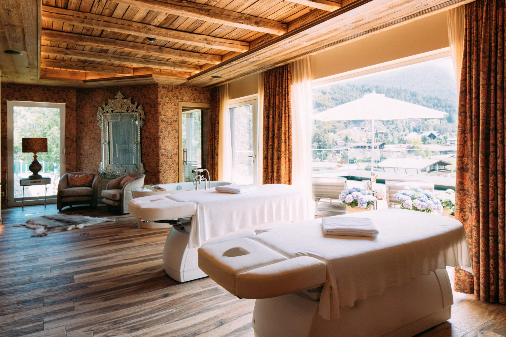 Zwei Massagebetten in einem stilvoll eingerichteten Raum mit Holzdecke und großen Fenstern, die den Blick auf eine sonnige Terrasse mit Blumen und Berglandschaft freigeben.