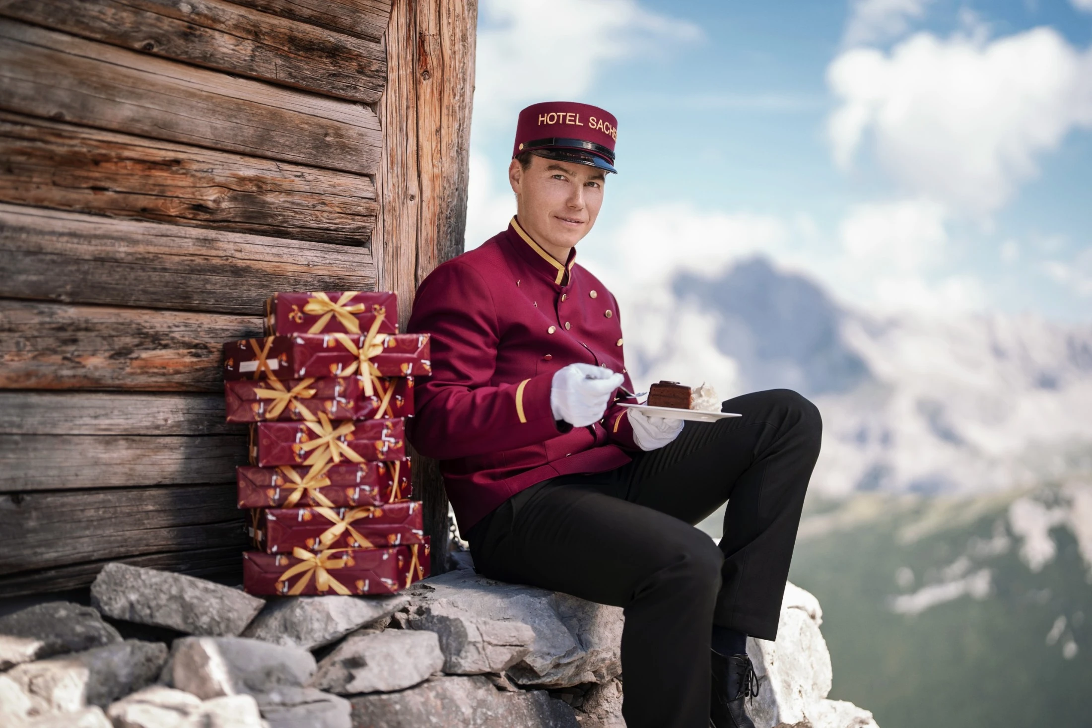 Ein Page des Hotel Sacher sitzt vor einer Almhütte mit Blick auf die Berge und genießt ein Stück Original Sacher-Torte. Neben ihm stapeln sich mehrere rot-goldene Schachteln der der Original Sacher-Torte.