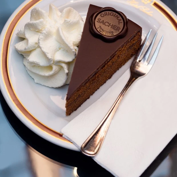 Ein Stück Original Sacher-Torte mit einer Portion Schlagobers auf einem weißen Teller mit Goldrand, dazu eine Gabel und eine Serviette.