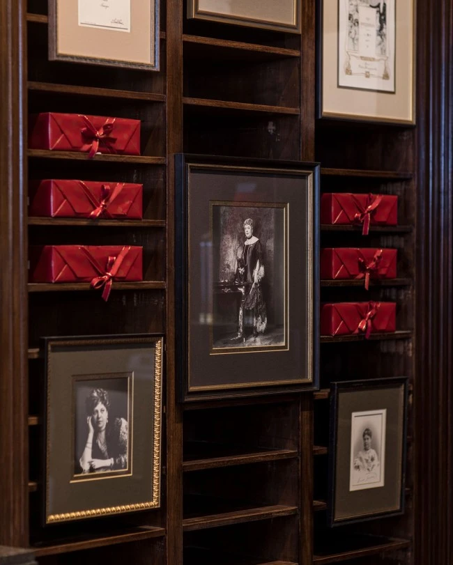 Holzwand mit integrierten Regalfächern, in denen mehrere rote Boxen mit der Original Sacher-Torte gestapelt sind. Dazwischen hängen gerahmte Schwarz-Weiß-Fotografien von Anna Sacher und anderen historischen Persönlichkeiten.