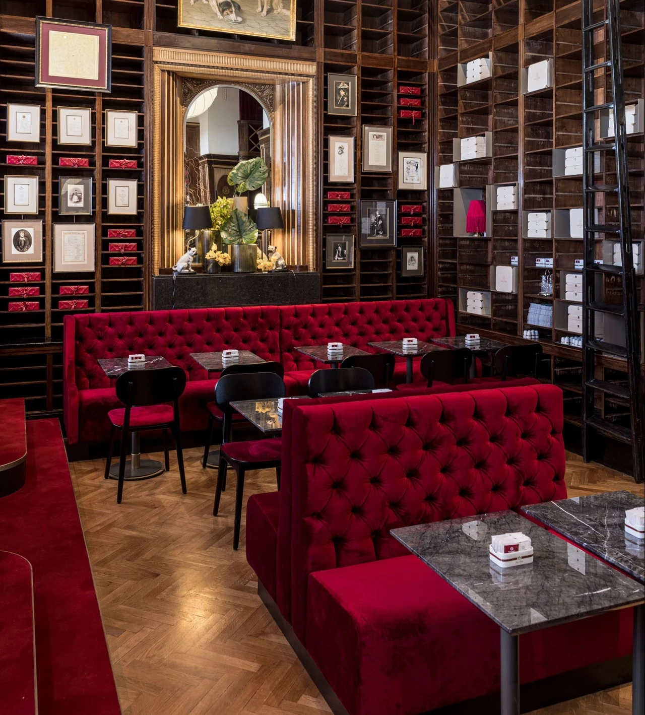 Der Raum zeigt eine elegante Café-Atmosphäre mit roten, samtbezogenen Sitzbänken und schwarzen Tischen mit Marmormuster. Die Wände sind von hohen Holzregalen eingerahmt, in denen teils rote, teils weiße Sacher-Holzschachteln und gerahmte Bilder dekorativ angeordnet sind. In der Mitte der Rückwand steht ein Spiegel mit vergoldetem Rahmen, flankiert von zwei schwarzen Lampen mit goldenen Elementen und floralen Dekorationen. Auf der rechten Seite des Raumes führt eine schwarze Leiter an den hohen Regalen entlang.