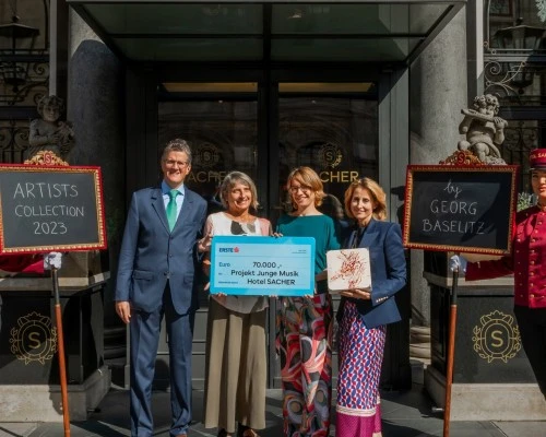 Vor dem Eingang des Hotel Sacher stehen vier Personen, darunter Alexandra Winkler (rechts) und Georg Gürtler (links), und präsentieren gemeinsam einen symbolischen Scheck über 70.000 Euro, der an das Projekt Junge Musik geht. Die Summe wurde durch die Artists Collection 2023 gesammelt, wie auf der linken Tafel zu lesen ist. Auf der rechten Tafel ist der Name des Künstlers Georg Baselitz vermerkt. Alexandra Winkler hält außerdem eine der gestalteten Sacher-Holzschachteln in den Händen.