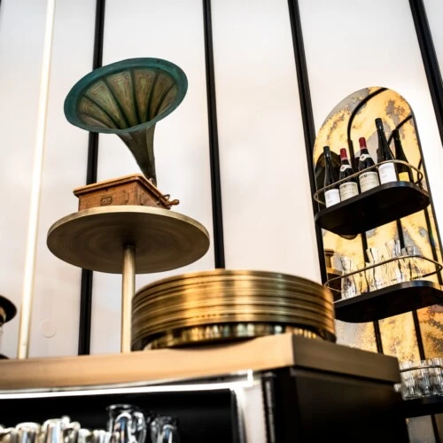 Ein stilvoll eingerichteter Barbereich mit einem auffälligen Grammophon auf einem Podest. Im Vordergrund sind Gläser und goldene Tabletts zu sehen. An der Rückwand befinden sich Regale mit verschiedenen Flaschen Wein und Spirituosen sowie weiteren Gläsern. Die Wand ist mit goldfarbenen Spiegel-Elementen dekoriert, die dem Raum eine elegante Note verleihen.
