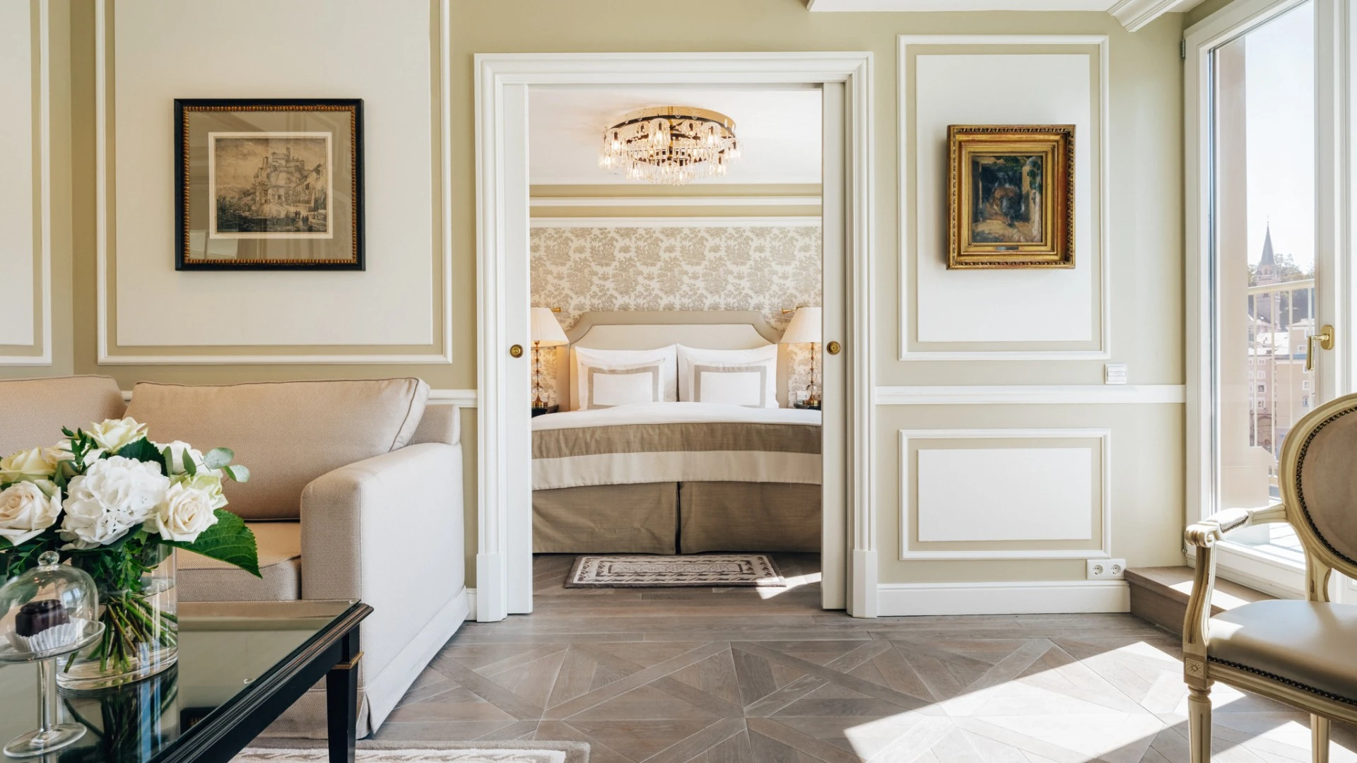 Ein elegantes Wohnzimmer mit einem beigen Sofa auf der linken Seite, daneben ein Couchtisch mit Glasplatte, auf dem ein Blumenstrauß und zwei kleine Glashauben mit Original Sacher Würfel stehen. Durch eine offene Tür ist ein Schlafzimmer mit einem großen Bett sichtbar, das von zwei Nachttischen mit Lampen eingerahmt wird. An den Wänden hängen gerahmte Bilder, die Farben des Raumes sind in warmen Creme- und Beigetönen gehalten.