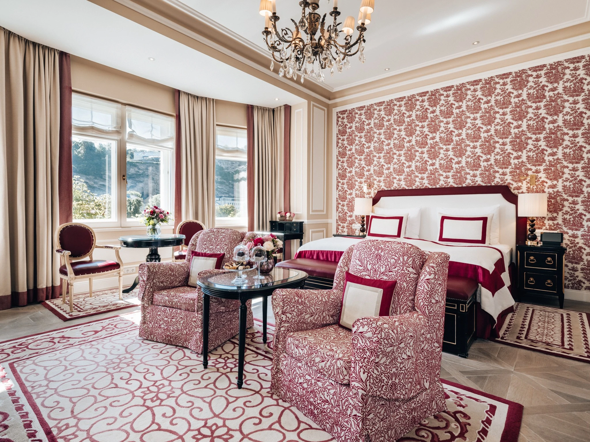 Zwei gemütliche Sessel mit floralem Muster und passenden Kissen stehen auf einem Teppich mit rot-weißem Ornamentmuster. Davor ein Glastisch mit einer Etagere, auf der sich verschiedene Süßigkeiten und zwei Original Sacher Würfel unter Glashauben befinden. Im Hintergrund ein großes Bett mit rotem Bettrahmen und abgestimmter Bettwäsche. Neben dem Fenster steht ein kleiner runder Tisch mit einem Blumenstrauß und zwei eleganten Stühlen. Die großen Fenster lassen viel Tageslicht in den Raum.