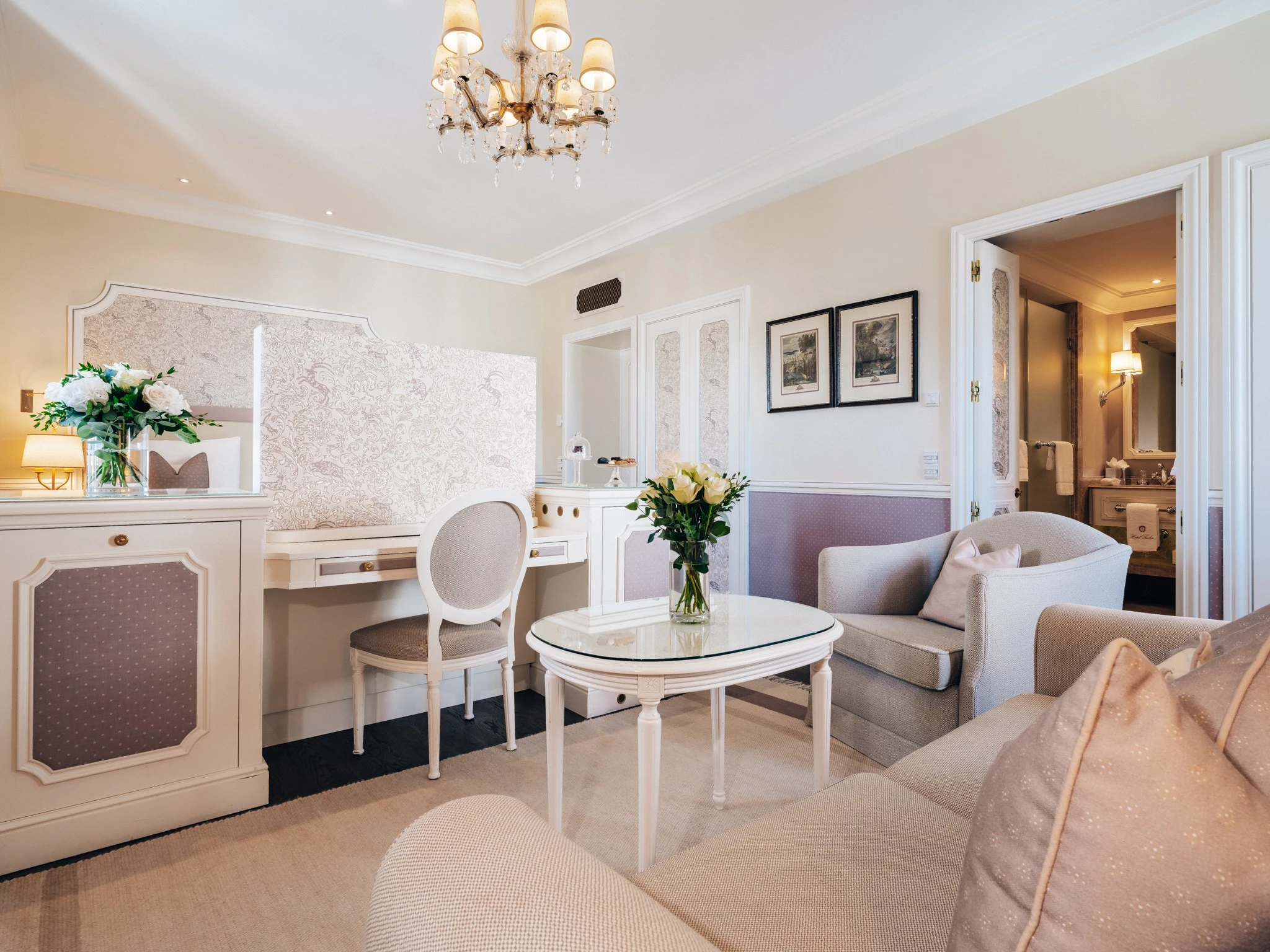 Ein heller Wohnbereich mit einem Sofa, einem Sessel und einem kleinen runden Tisch, auf dem ein Strauß weißer Rosen steht. Im Hintergrund ein Schreibtisch mit gepolsterter Stuhllehne, einer Tischlampe und weiteren Blumen. An der Wand hängen zwei gerahmte Bilder und durch eine geöffnete Tür ist ein Badezimmer sichtbar.