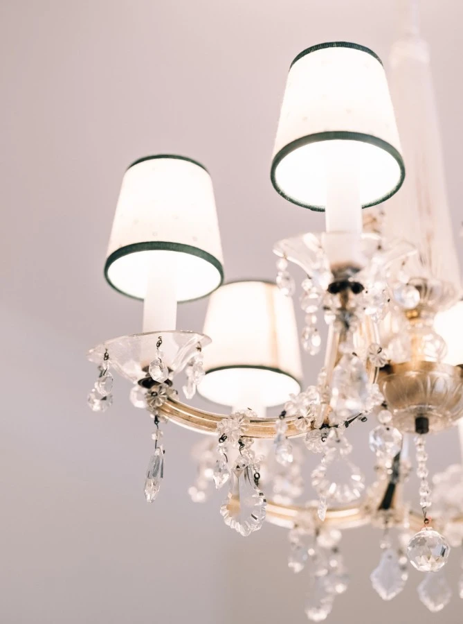 Ein Kronleuchter mit mehreren Leuchten, die mit kleinen weißen Lampenschirmen mit grünem Rand versehen sind. Der Leuchter ist mit zahlreichen funkelnden Kristallen geschmückt, die elegant von den Armen des Leuchters herabhängen.