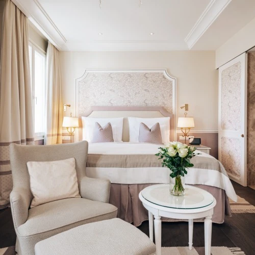 Ein gemütliches Schlafzimmer mit einem großen Bett, das mit hellen Kissen und einer dezenten Tagesdecke ausgestattet ist. Vor dem Bett befinden sich ein Sessel mit Hocker sowie ein runder Beistelltisch mit einem Blumenstrauß. Der Raum ist in sanften Beige- und Rosatönen gehalten, die Tapete hinter dem Bett ist dezent gemustert. Rechts führt eine Tür in das angrenzende Badezimmer.