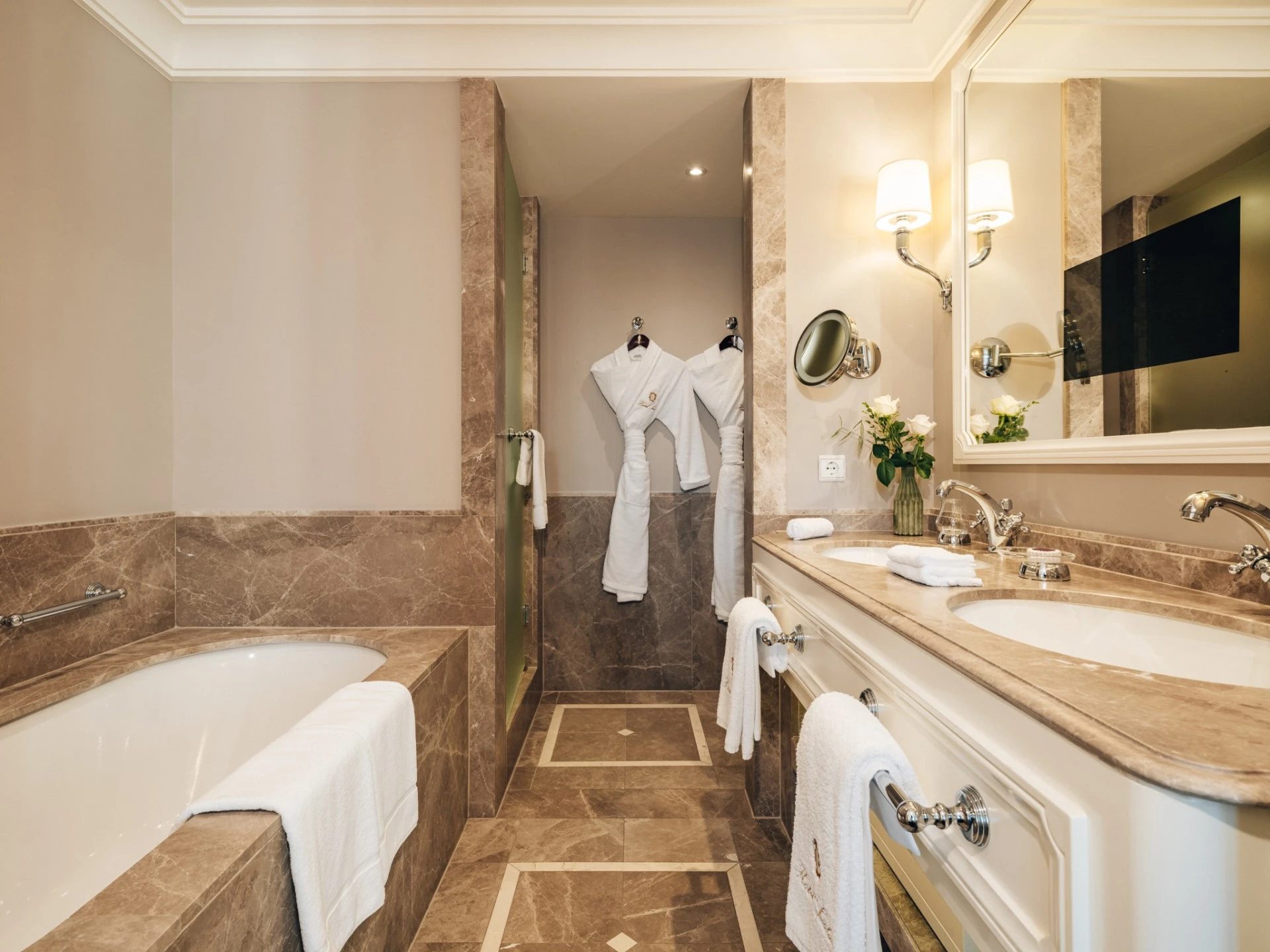 Ein Badezimmer mit einer in Marmor eingefassten Badewanne, einem Doppelwaschbecken mit Spiegel und zwei Bademänteln auf Kleiderbügeln.