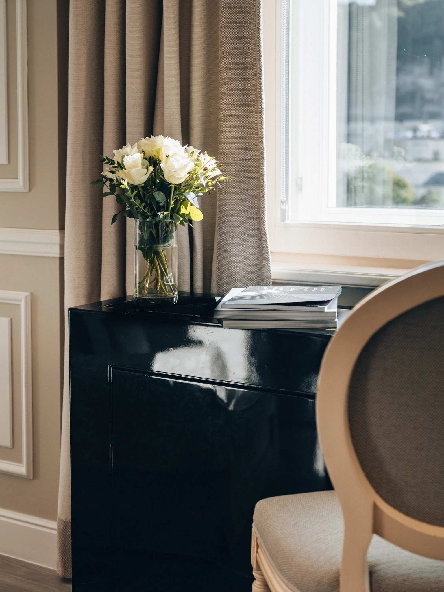 Ein schwarzes Sideboard mit einem Blumenstrauß aus weißen Rosen darauf. Daneben liegen zwei Bücher. Im Vordergrund ein Polsterstuhl, im Hintergrund helle Vorhänge und ein großes Fenster.